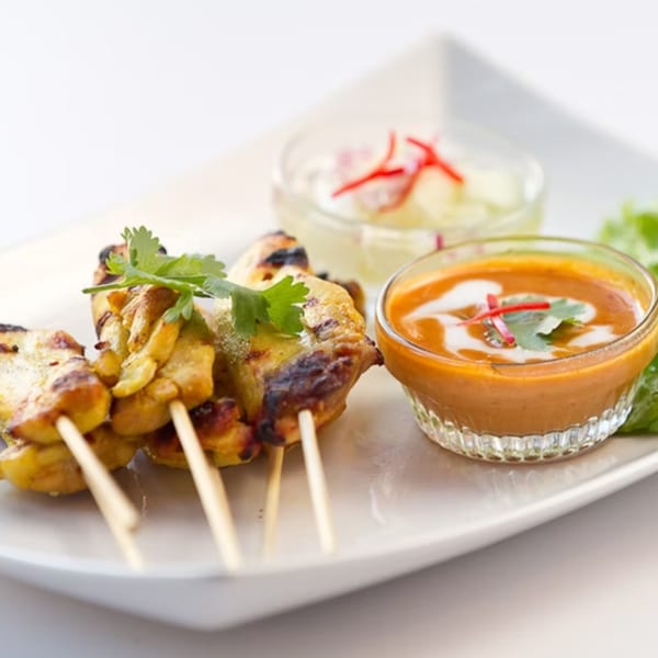 Grilled chicken satay skewers with peanut sauce and cucumber relish on a white plate.
