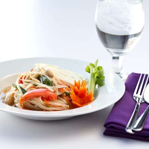A Thai papaya salad with peanuts, tomatoes, and a carved carrot garnish, next to a water glass.