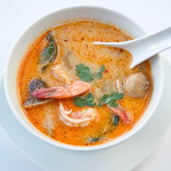 A white bowl of vibrant orange Tom Yum soup with shrimp, mushrooms, and cilantro, with a spoon.