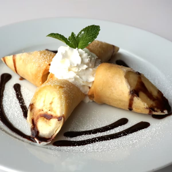 Four fried banana spring rolls with whipped cream, mint, chocolate sauce, and powdered sugar.