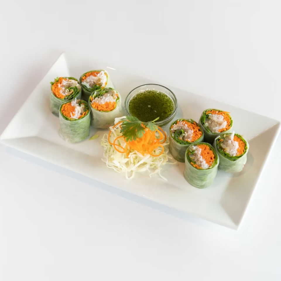 Eight fresh Thai spring rolls with shredded carrots, lettuce, and white filling, served with green dipping sauce.